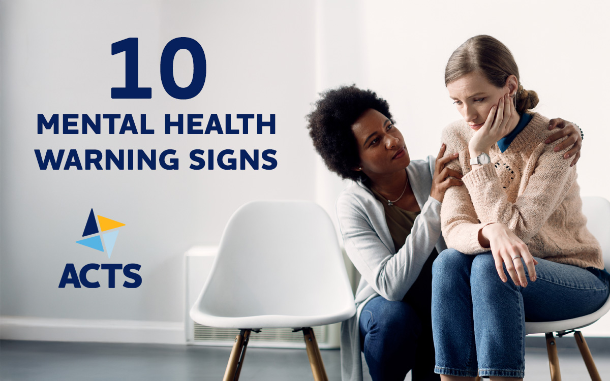 A supportive woman gently comforts a distressed woman sitting beside her, who appears sad and withdrawn. To the left, bold text reads "10 Mental Health Warning Signs" above the ACTS logo, which features blue and yellow triangular shapes. The setting is a simple, bright room with white chairs.