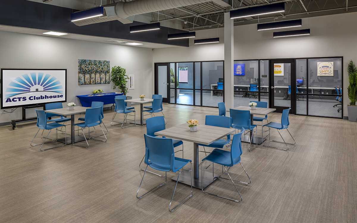 A modern, brightly lit community space labeled “ACTS Clubhouse” with several square tables and blue chairs arranged for seating. A large screen on the left displays the ACTS Clubhouse logo. The room features light wood flooring, potted plants, and wall art. In the background, there are glass-walled rooms with computer workstations and motivational posters, suggesting a multi-use facility for gatherings, meetings, or computer access.